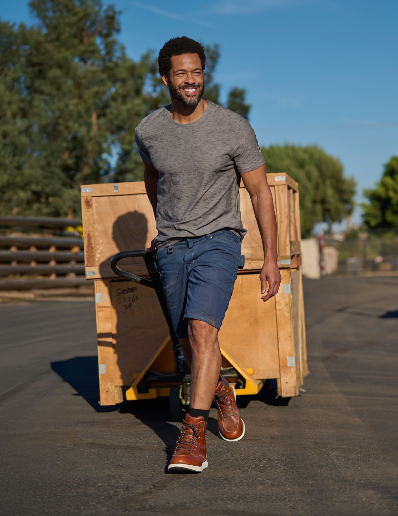 Man in blue shorts pushing a pallet truck with wooden pallets outdoors on a sunny day.