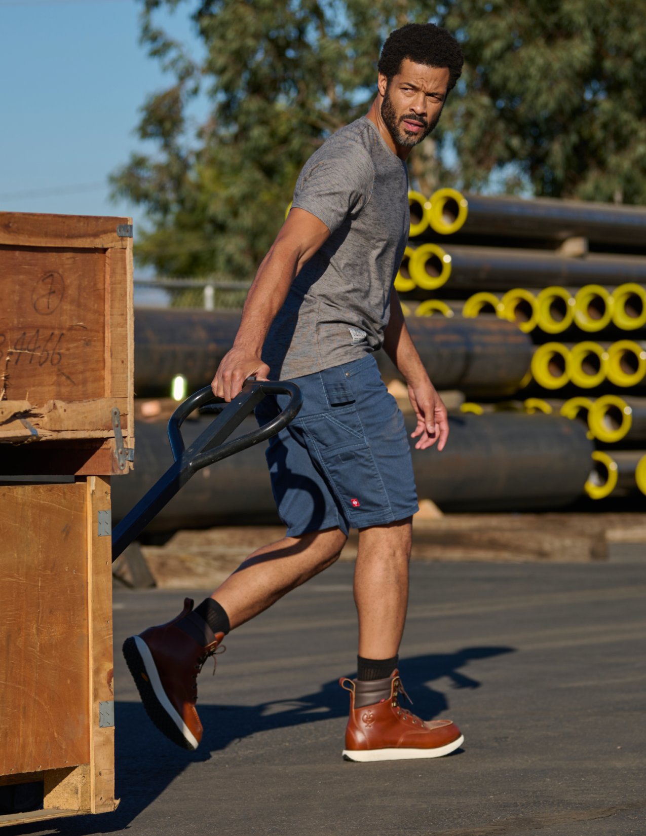 Man in gray t-shirt and blue shorts walking outdoors with pipes and wooden crates in the background