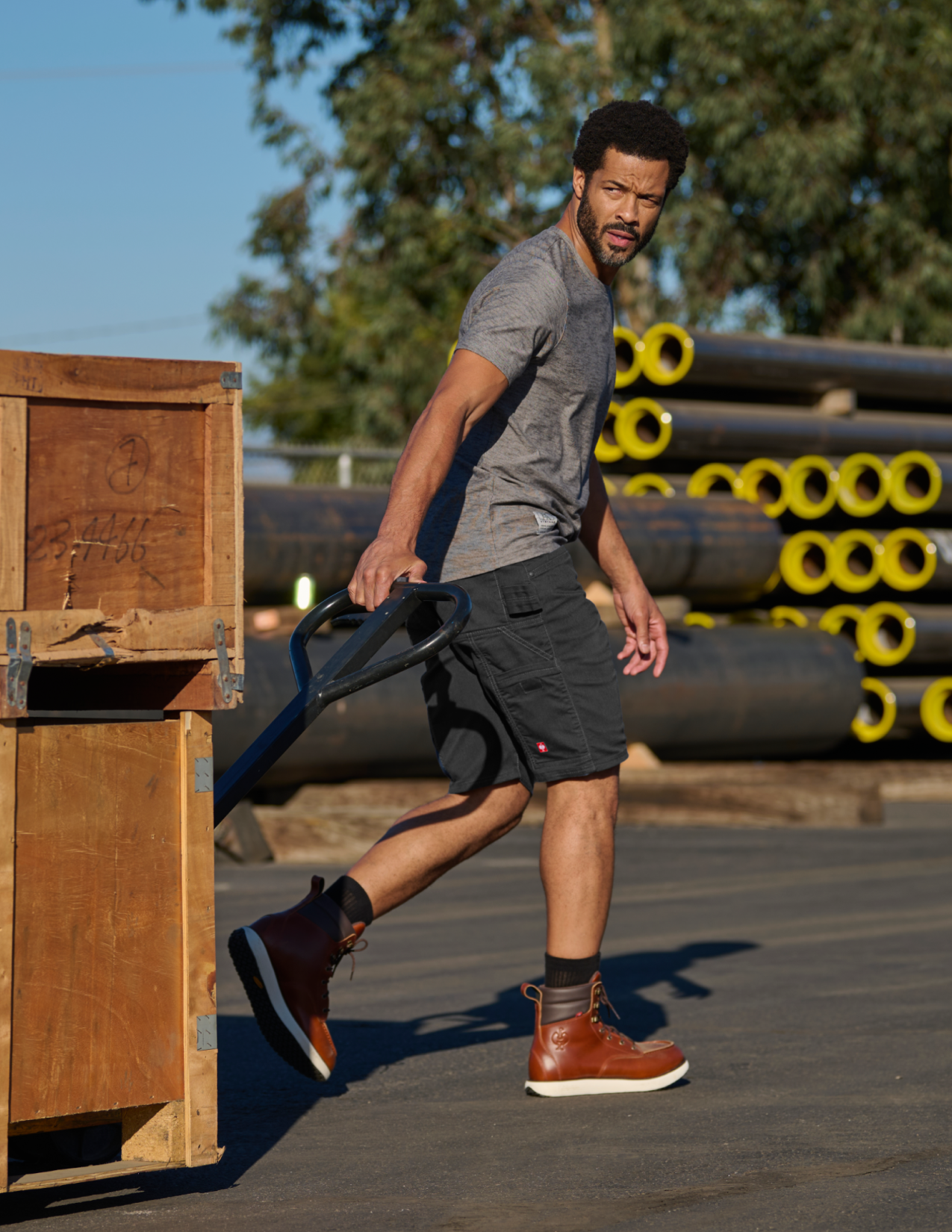 Man in black shorts lifting crates outdoors with pipes in the background