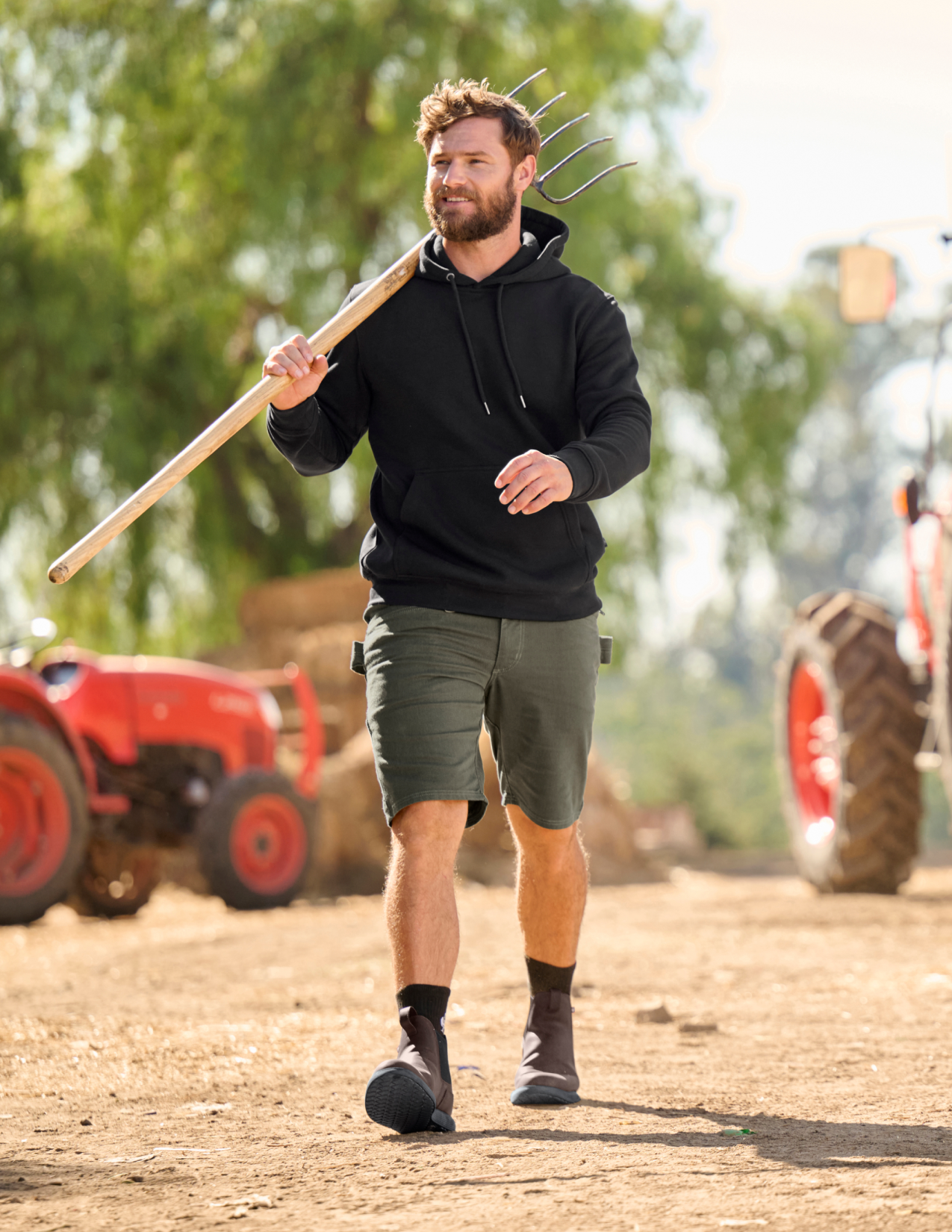 Man walking outdoors holding a pitchfork with tractors in the background