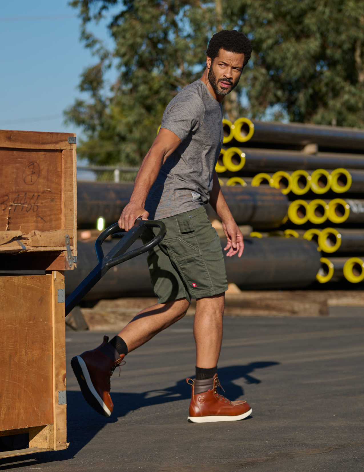 Man wearing green shorts pushing a wooden crate with stacks of pipes in the background.