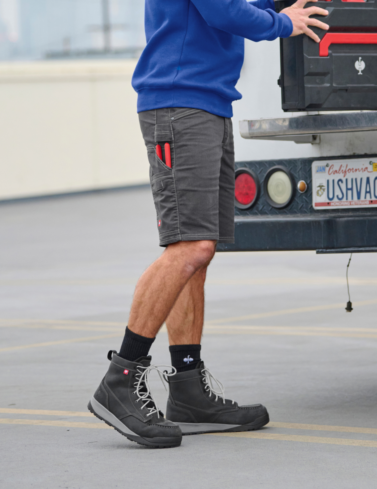 Person wearing gray shorts with red accents, black boots, and a blue sweatshirt, standing next to a truck.