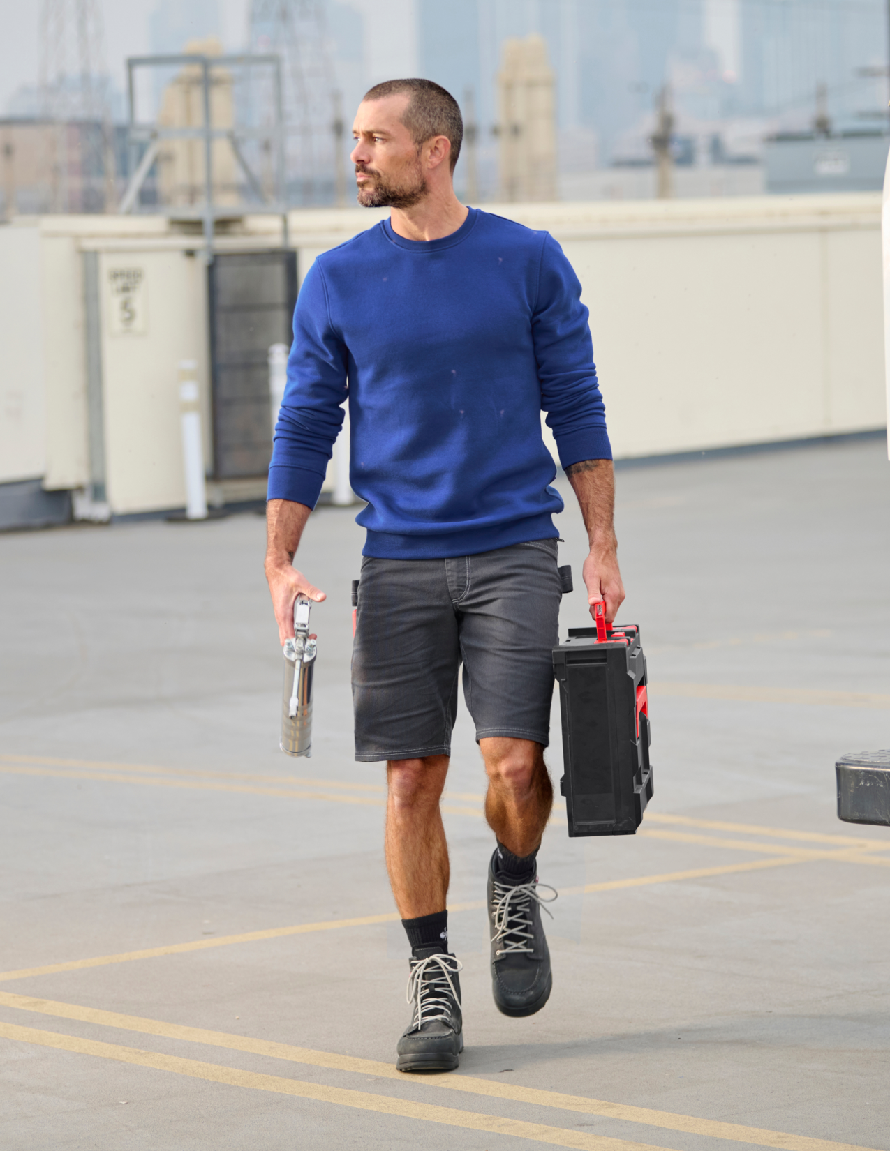 Man in blue sweater and shorts carrying tools on a rooftop
