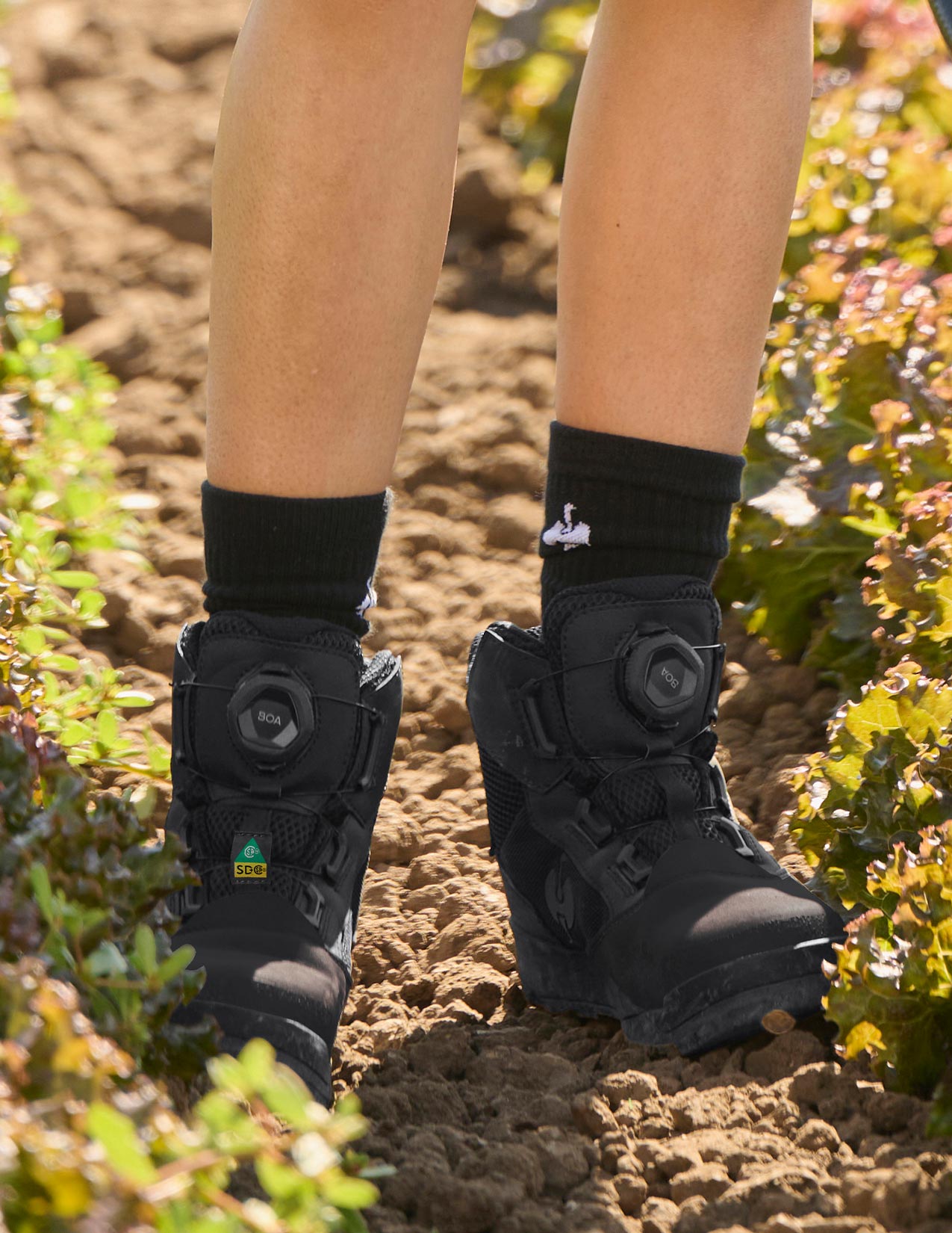 Person wearing black work boots with visible brand logo on a dirt ground.