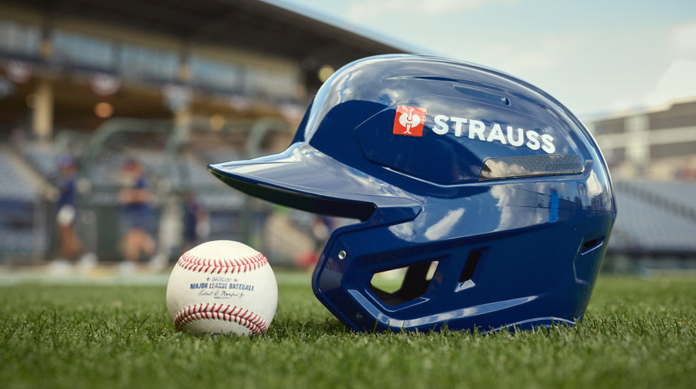 A blue Strauss baseball batting helmet sits on green grass next to a baseball on a baseball field, with a blurred stadium background.