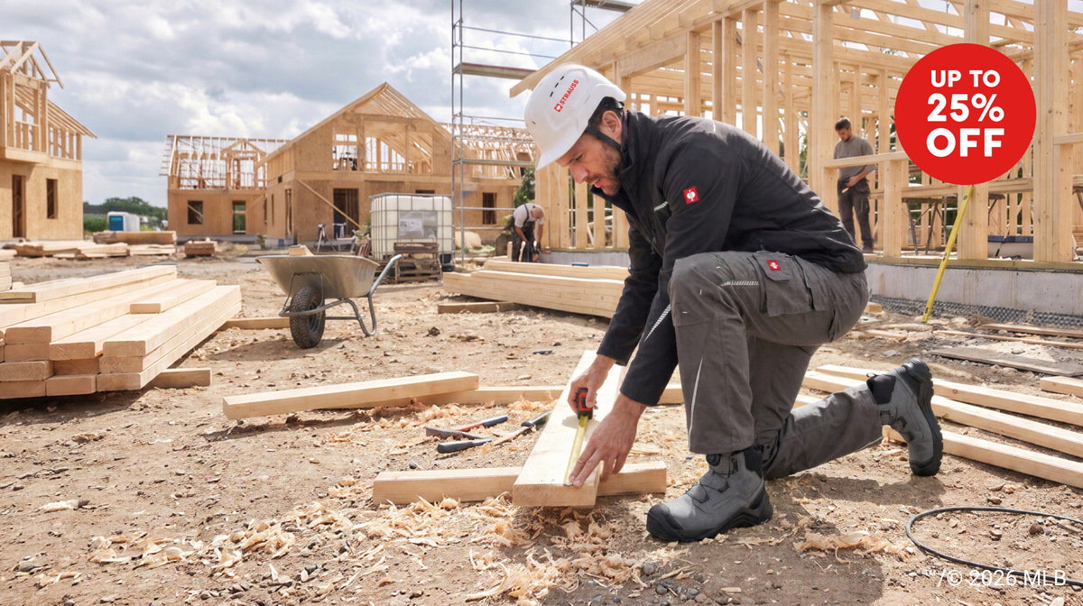 A man kneeling down to take measurement on a wooden board. The man is wearing black and grey STRAUSS workwear, a white helmet and gray work boots.