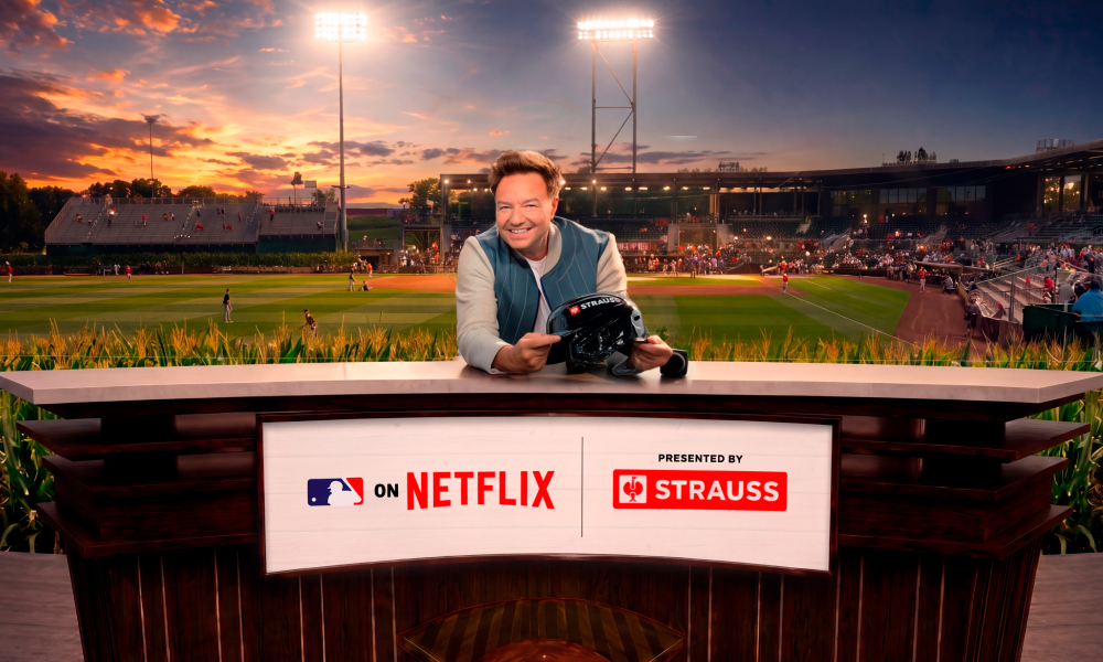 Henning Strauss sits behind a desk in a studio setup at Field of Dreams, holding a black baseball helmet. The background shows a baseball stadium at sunset with players on the field and crowd seating.