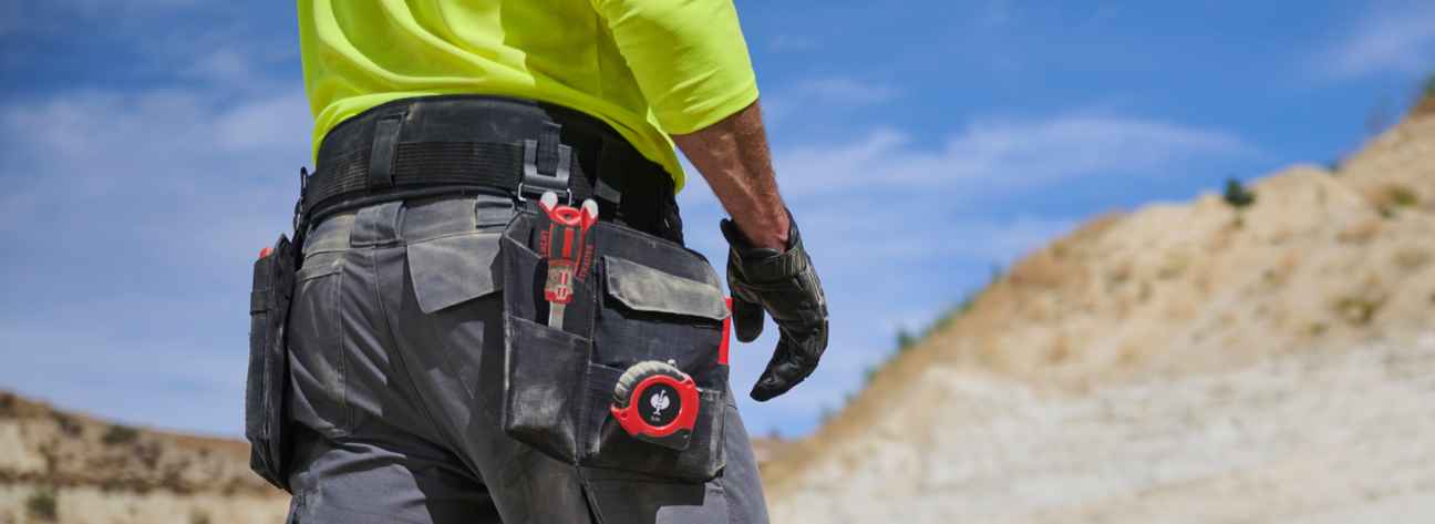 A worker wearing a neon yellow shirt and gray cargo pants has red tools attached to their black work belt at an outdoor construction site.