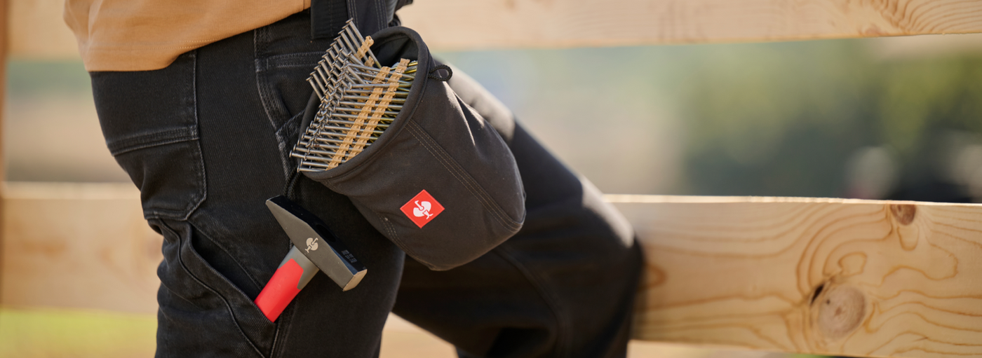 A worker wearing black cargo pants has a gray tool pouch with nails and a hammer attached to their belt.