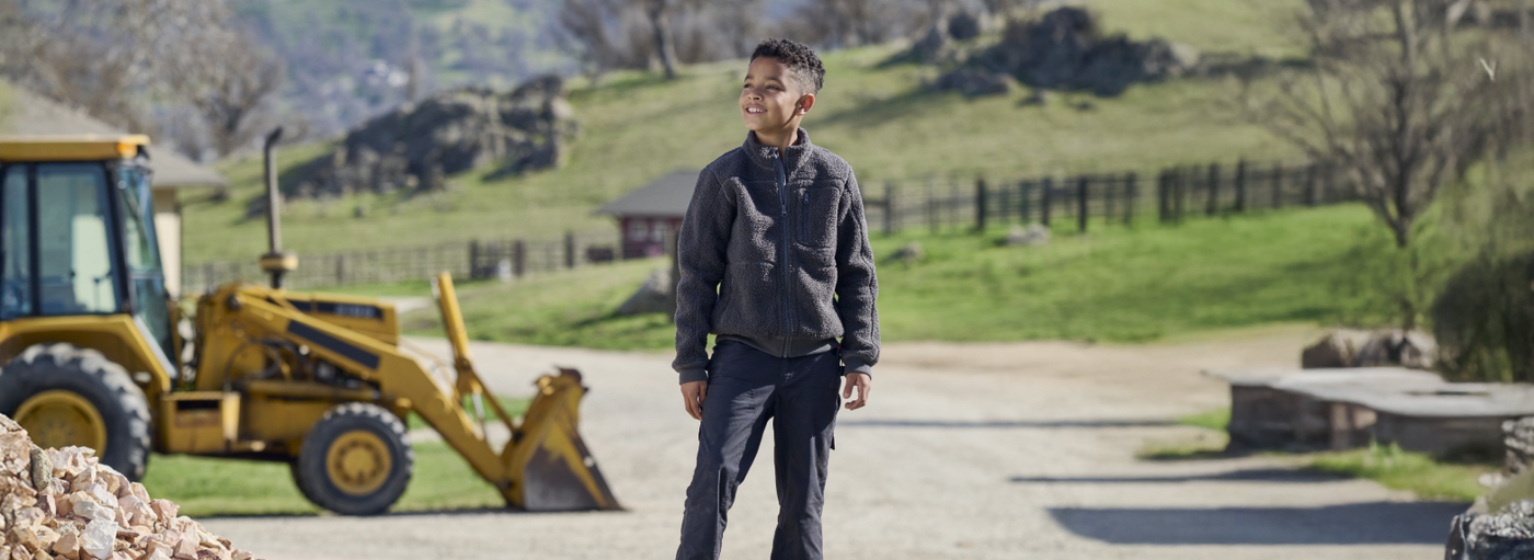Child wearing a dark fleece jacket and work pants on rural construction site with tractor.