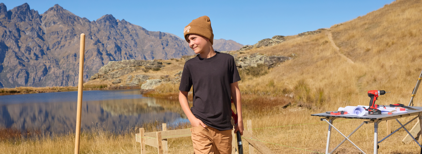A boy stands outdoors near a lake and mountains, with construction tools on a table nearby.