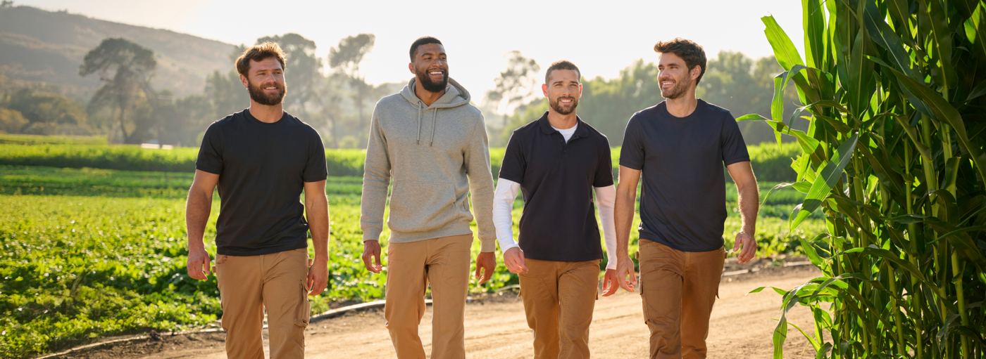Four men dressed in casual attire walking along a dirt path, surrounded by lush greenery and sunlight filtering through trees.