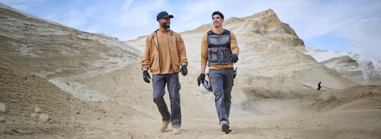 Two men wearing tan jackets and gray cargo pants are walking through a rocky outdoor landscape with one wearing a tool vest.