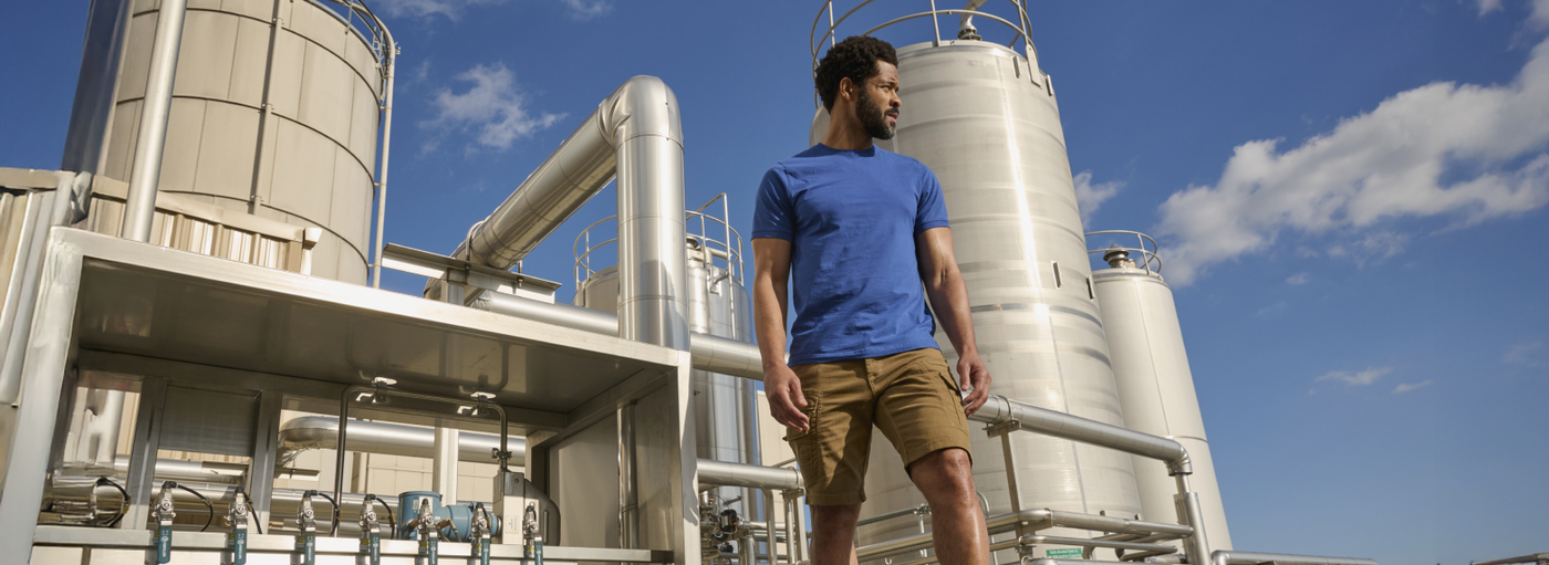 A man wearing a blue t-shirt and tan cargo shorts is standing at an industrial facility with white storage tanks and metal structures.