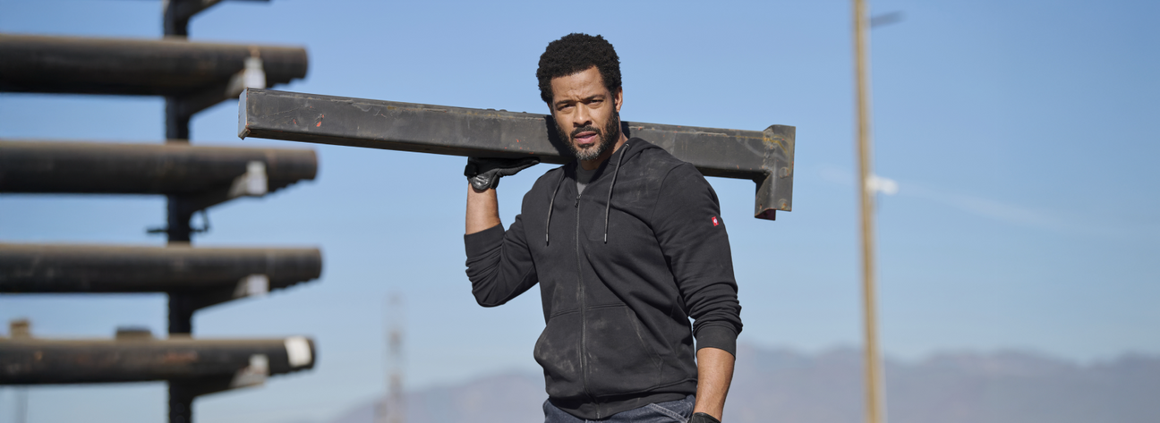 A man wearing a dark gray full-zip hoodie and jeans is carrying a metal beam on his shoulder at an outdoor construction site with wooden materials and blue sky.
