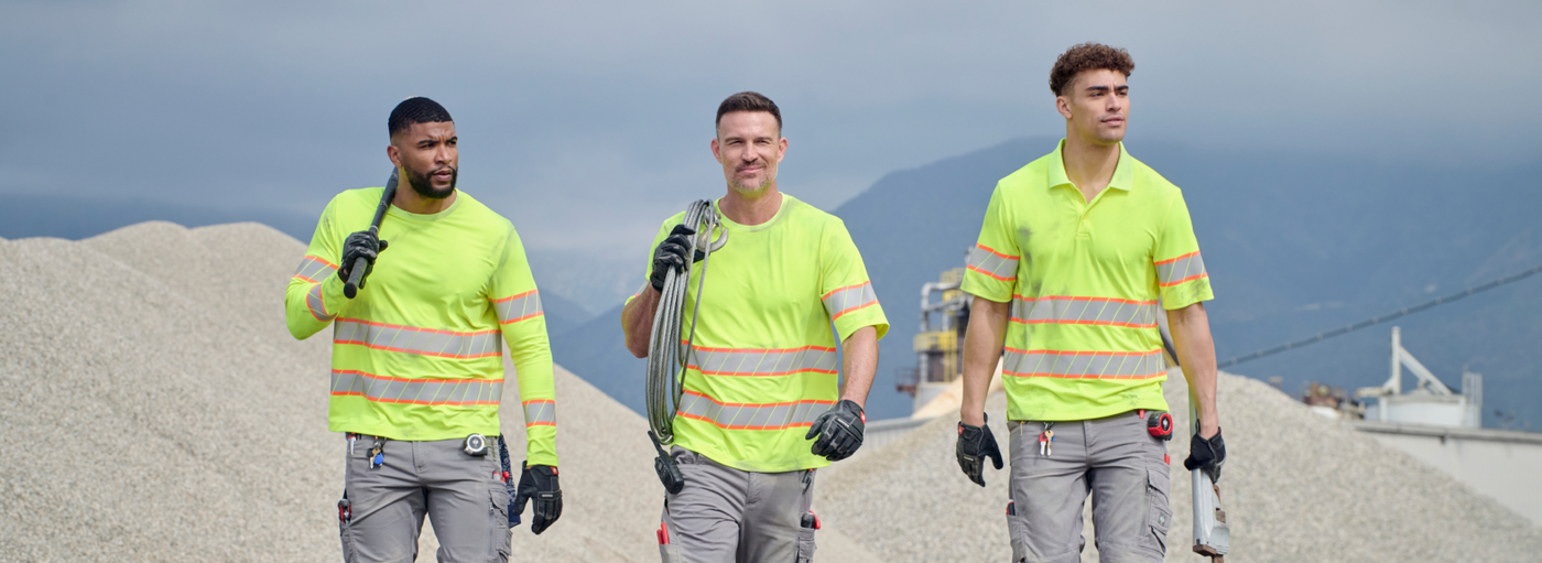 Three workers wearing neon yellow high visibility shirts with reflective stripes and gray cargo pants are walking at an outdoor gravel site with mountains in the background.