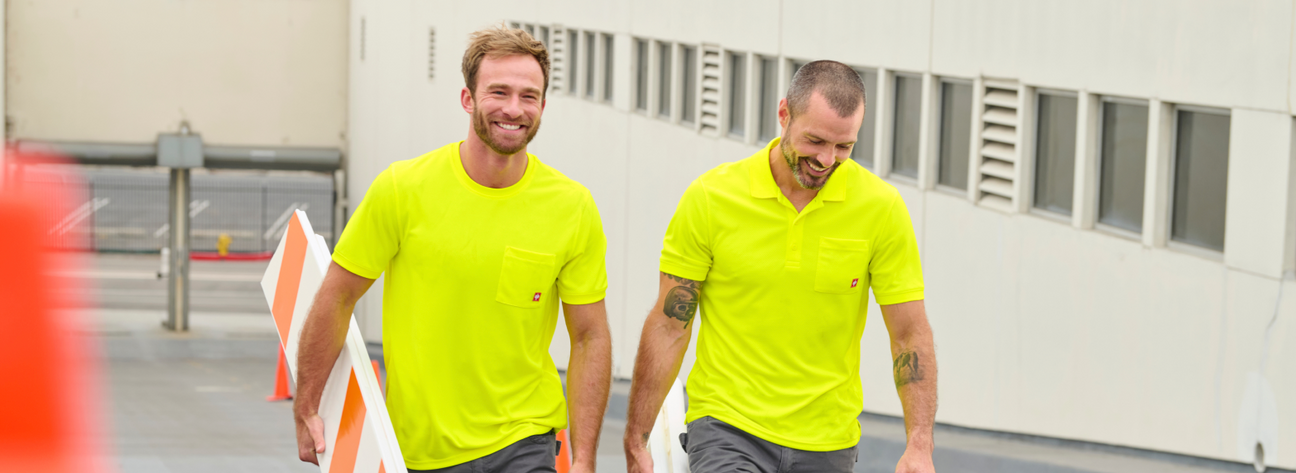 Two workers wearing neon yellow high visibility t-shirts and gray cargo pants are walking outdoors at an industrial facility .