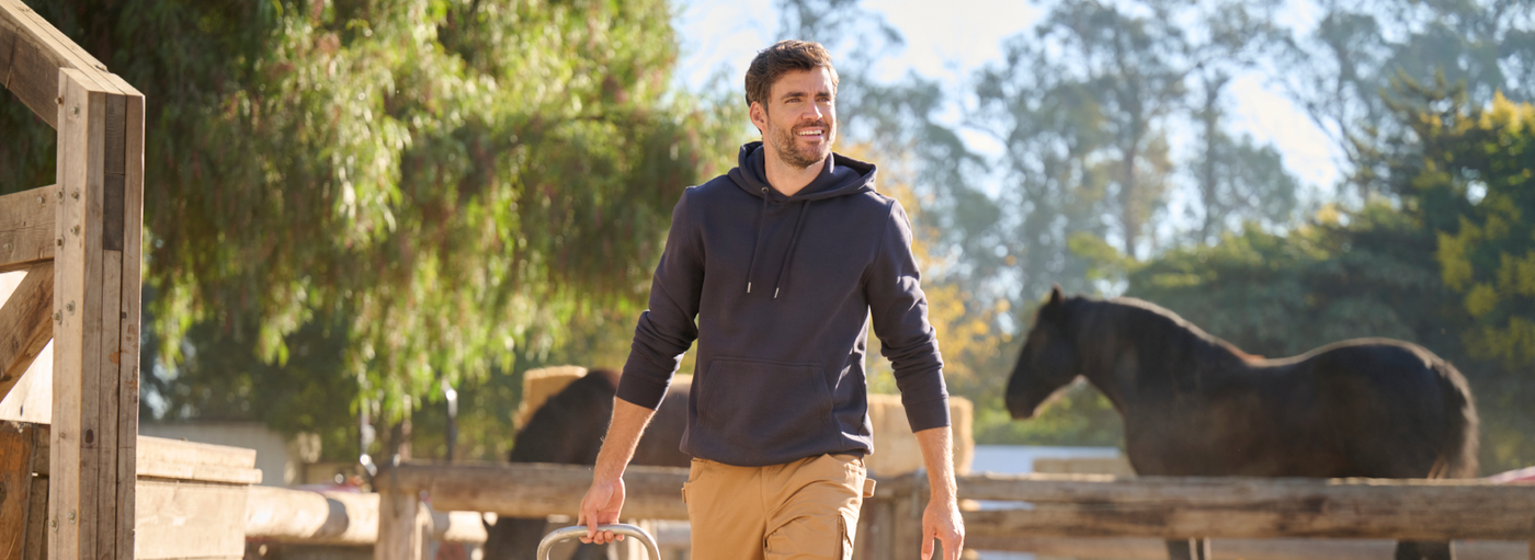 A man wearing a navy blue hoodie and tan cargo pants is walking at a rural farm with horses and wooden fences.