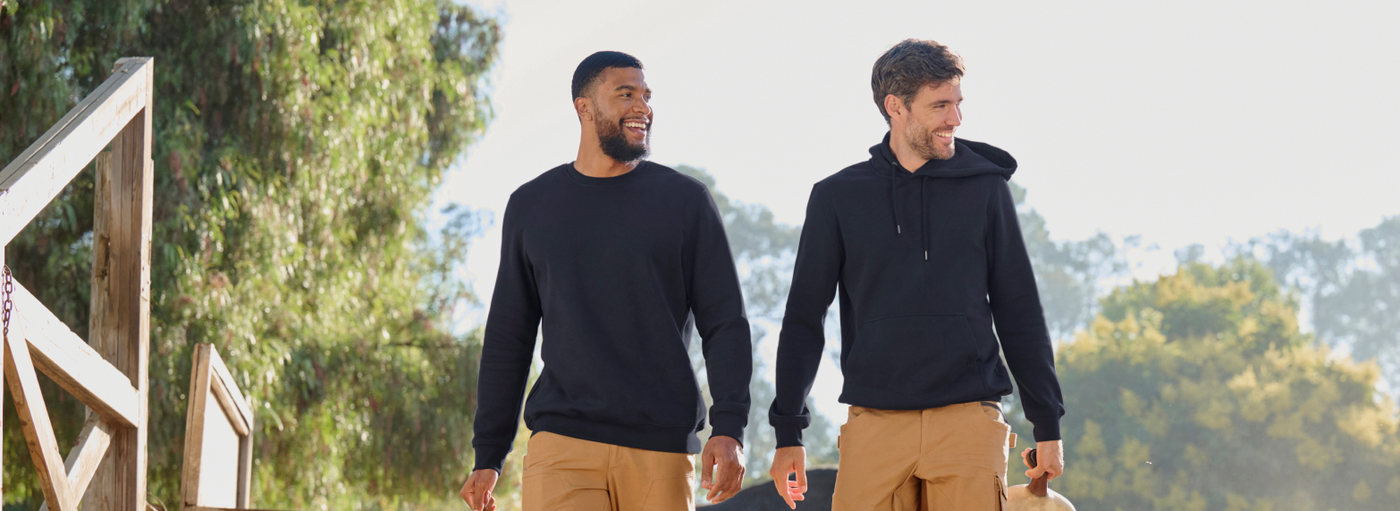 Two men wearing black sweatshirts and tan cargo pants are walking outdoors on a wooden deck with a scenic mountain landscape.