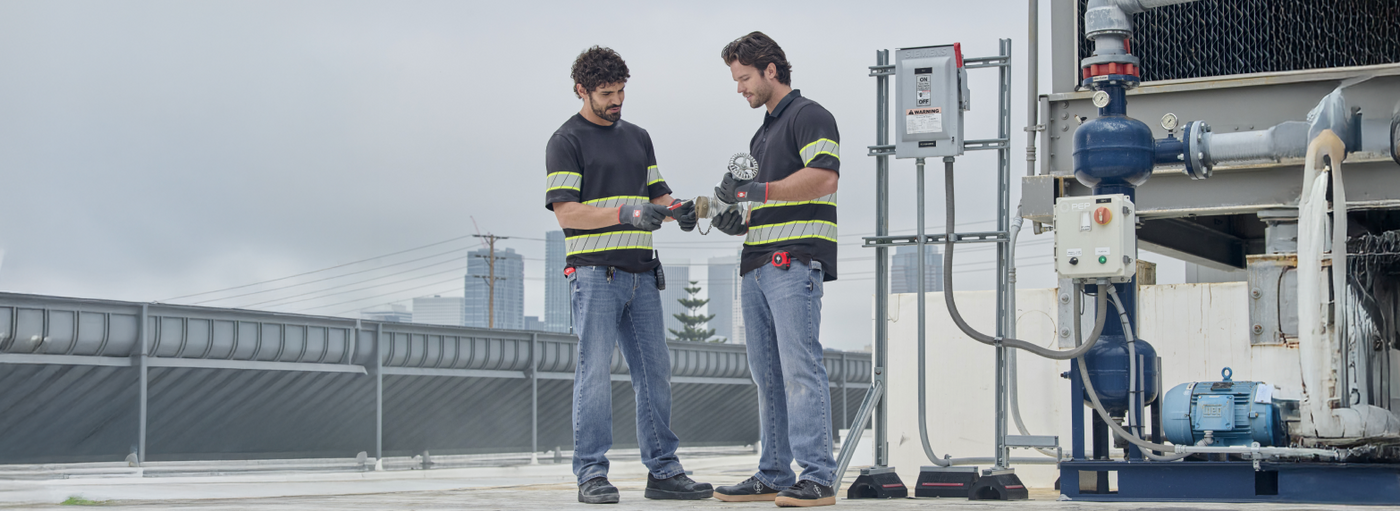 Two workers wearing black and yellow reflective safety shirts with blue jeans are standing on a rooftop with industrial equipment and city buildings in the background.