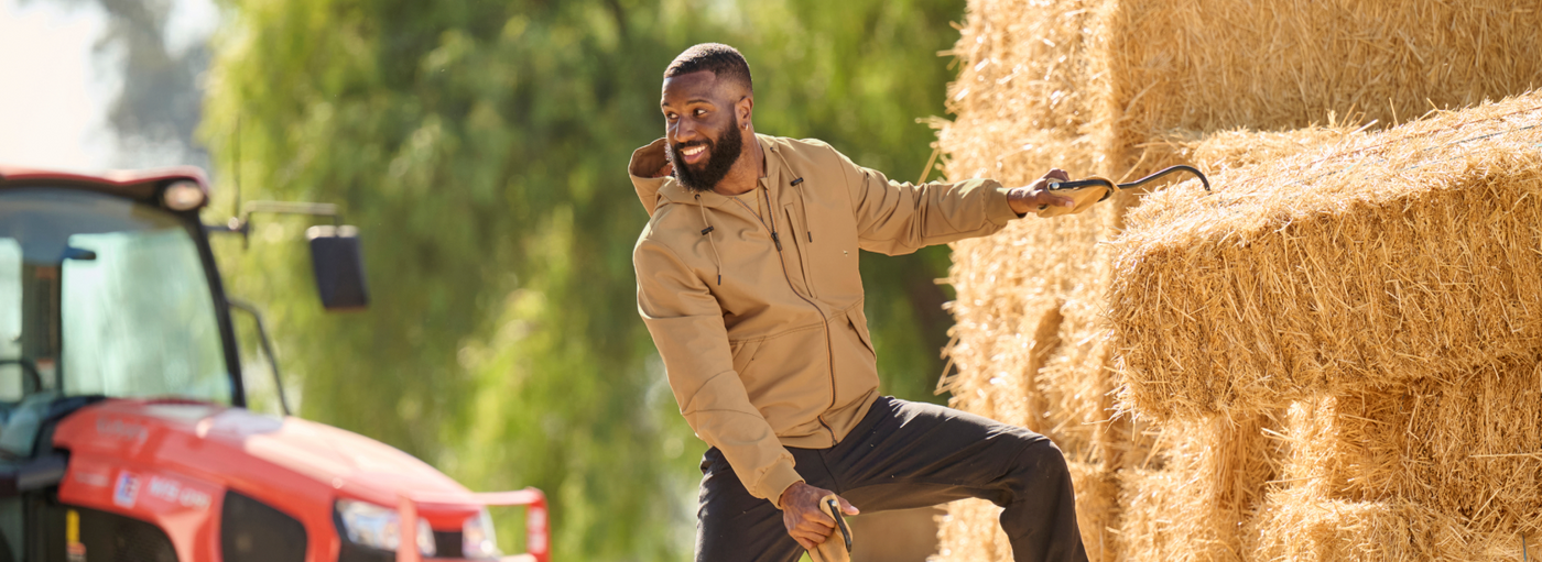A man wearing a tan work jacket is sitting on hay bales near a red tractor on a farm.