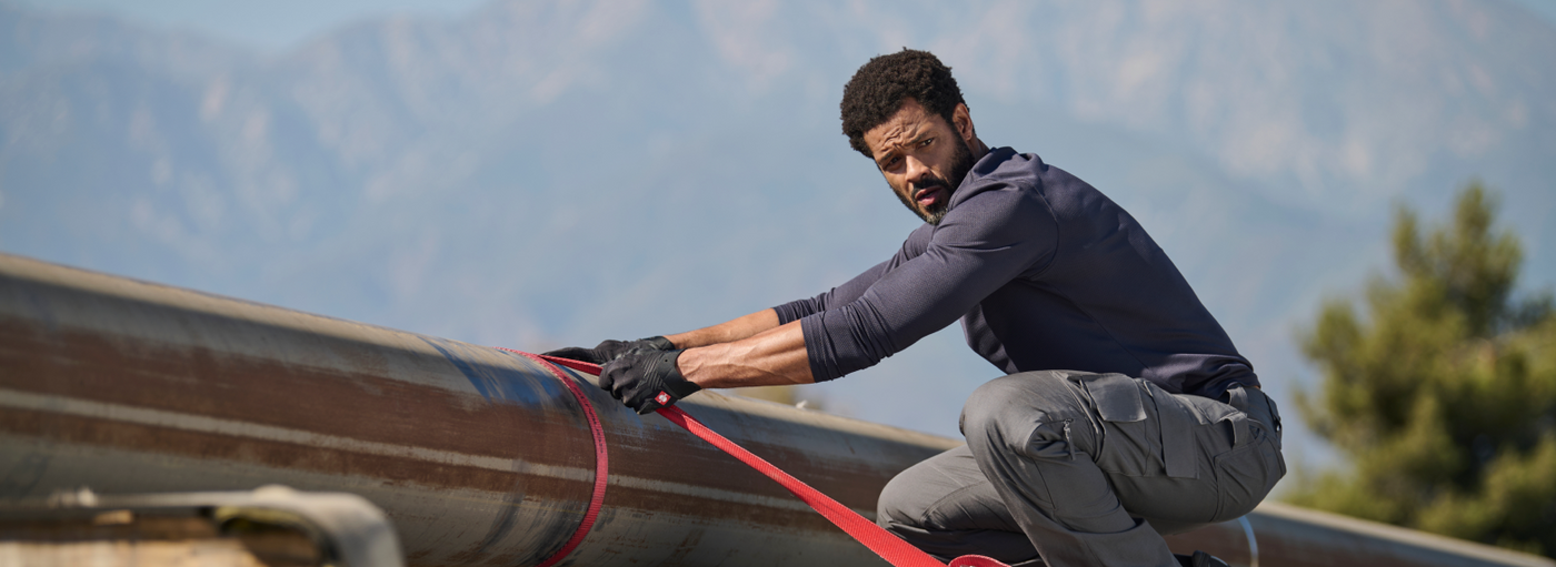 A man wearing a navy blue long-sleeve performance shirt and gray cargo pants  is securing a red strap on a metal structure at an outdoor work site.