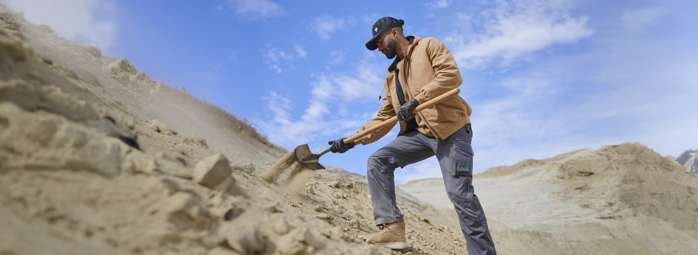 A worker wearing a tan work jacket, black baseball cap, and gray cargo pants with work gloves is digging with a shovel in sandy terrain with rocky hills.