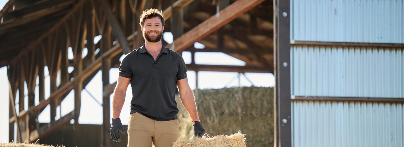 A man wearing a black polo shirt, tan work pants, and black work gloves is standing at a farm with a barn structure and hay bales.