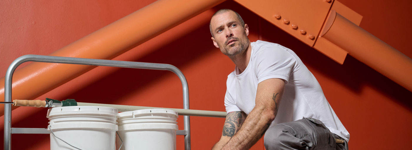 A man wearing a white t-shirt and gray cargo pants is kneeling with paint buckets at an orange industrial structure.