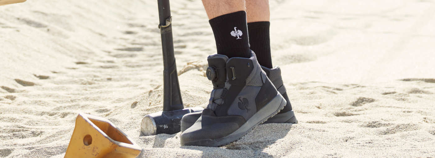 A person wearing black work boots with BOA lacing system and black crew socks is standing on sand with a shovel.