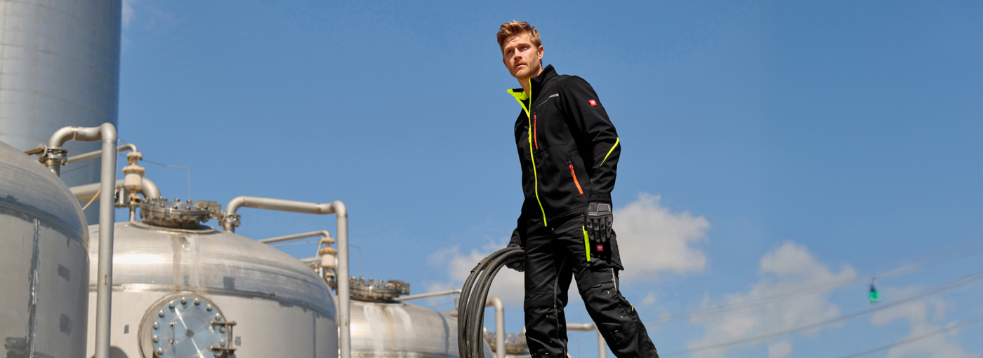 Person wearing a black and yellow jacket standing in front of industrial equipment with a blue sky background