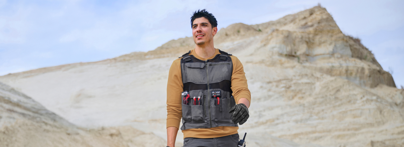 A man wearing a tan long-sleeve shirt, black tactical tool vest with multiple pockets and red-handled tools, and gray cargo pants is standing outdoors with rocky terrain in the background.