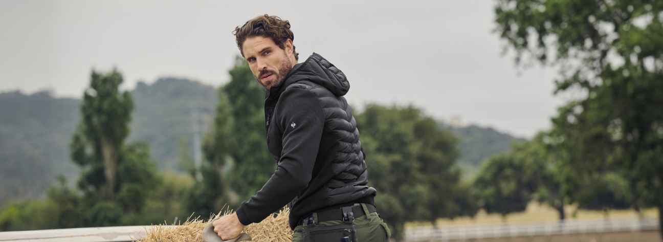 A man wearing a black quilted work jacket with hood and green cargo pants is leaning on hay bales at a rural farm with trees and mountains in the background.
