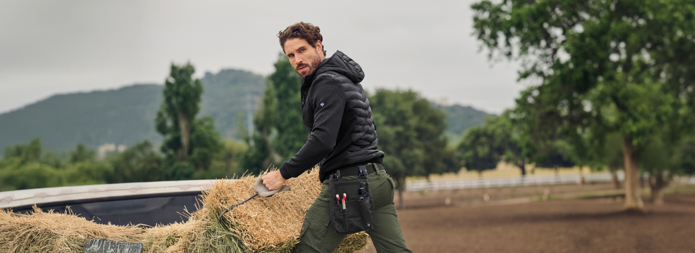 A man wearing a black hooded jacket and olive green work pants with tool pouches is loading hay bales on a rural farm.