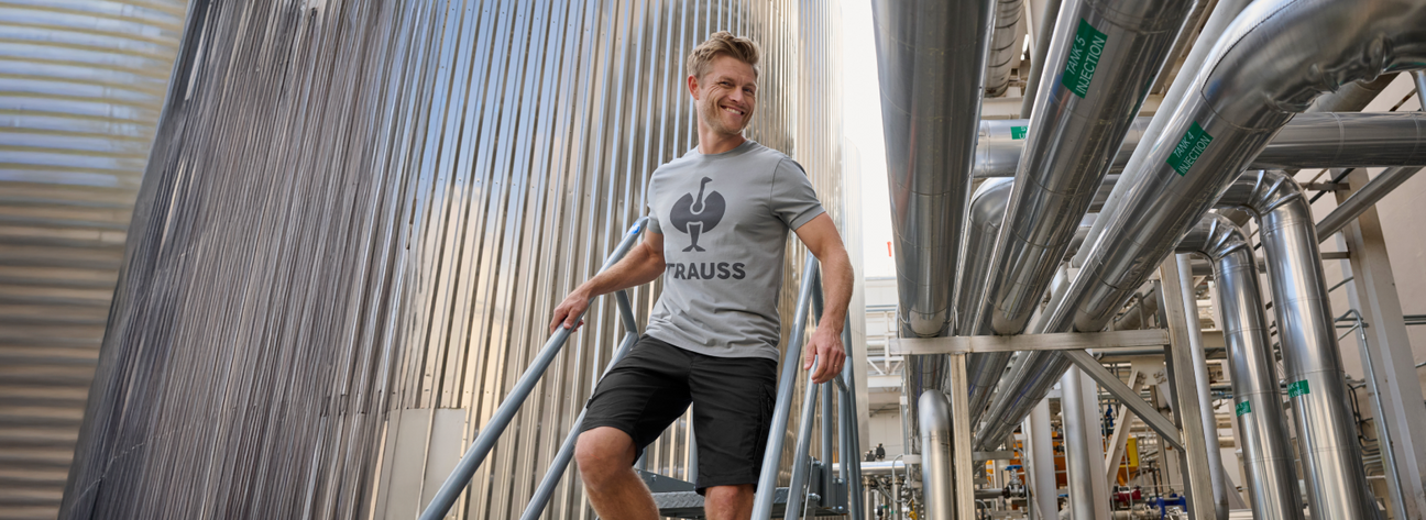 A man wearing a gray t-shirt and black cargo work shorts is standing on stairs at an industrial facility with steel tanks.