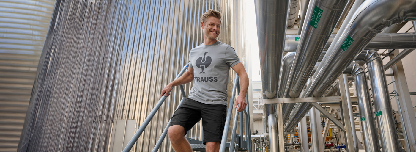 A man wearing a gray t-shirt and black cargo work shorts is standing on stairs at an industrial facility with steel tanks.