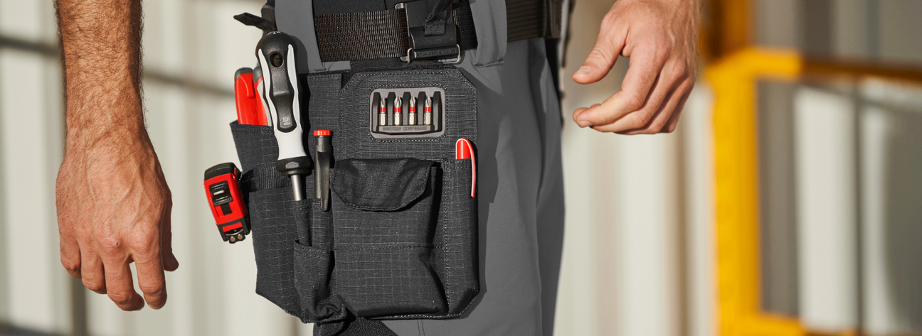 A worker wearing gray pants has a black tool bag with multiple screwdrivers and red tools attached to their waist.