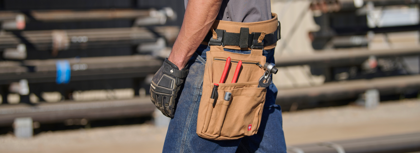 Worker wearing tan canvas tool belt with multiple pockets and tools at construction site.