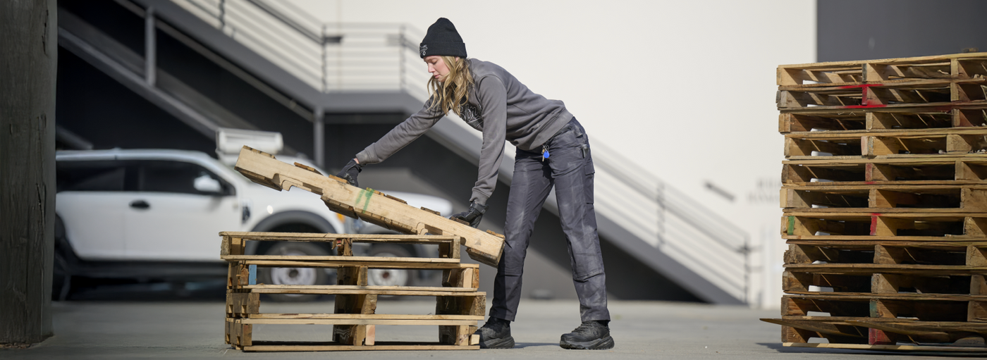 A woman wearing a gray hoodie and charcoal double front work pants is moving wooden pallets at a loading dock.