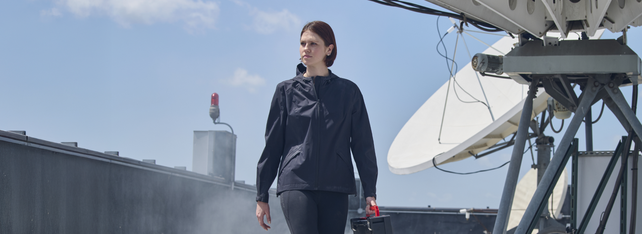 A woman wearing a navy blue lightweight jacket and dark pants is walking on a rooftop with industrial equipment.