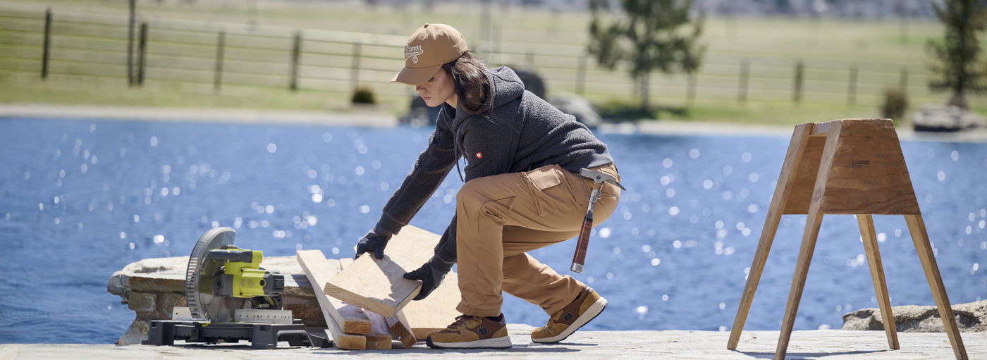 A woman wearing a tan baseball cap and khaki work pants is using a circular saw to cut wood near a lake.