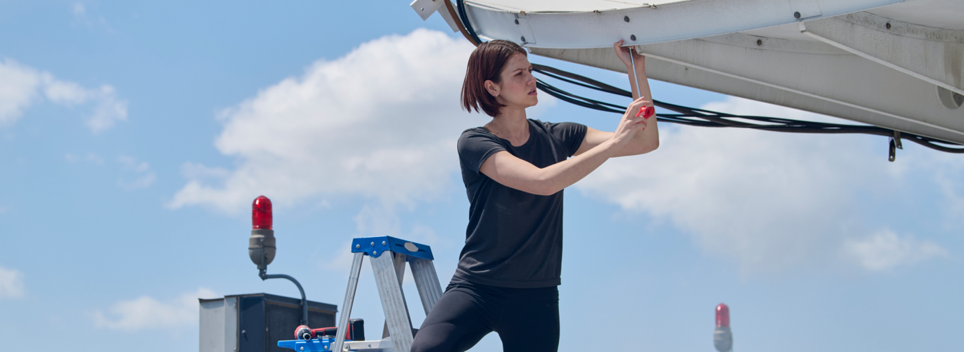 
A woman wearing a dark gray short-sleeve work shirt and black cargo pants is installing or adjusting equipment on a white industrial structure with a blue sky background.