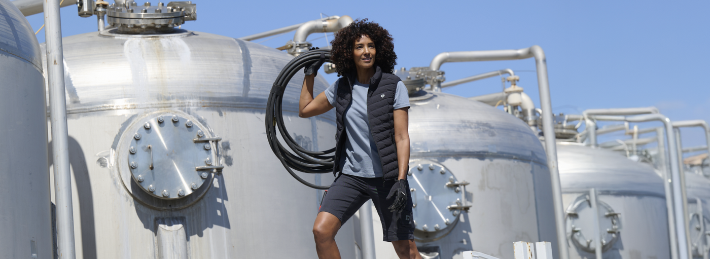 A woman wearing a navy puffer vest and black cargo shorts is carrying a coiled hose at an industrial facility with metal tanks.