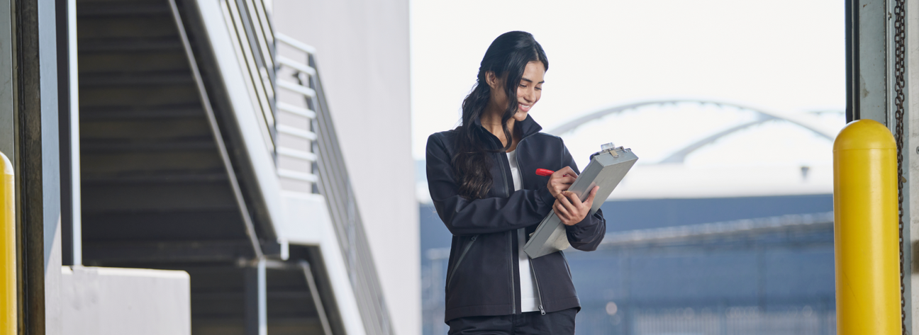 A woman wearing a black softshell jacket is writing on a clipboard at an outdoor loading dock.