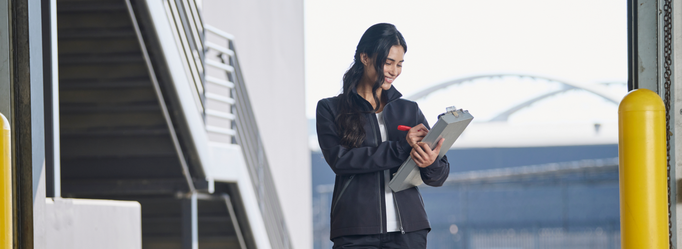 A woman wearing a black softshell jacket is writing on a clipboard at an outdoor loading dock.