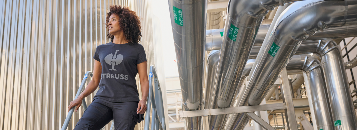 A woman wearing a navy blue graphic t-shirt and black cargo pants is standing on industrial stairs with metal tanks and pipes.