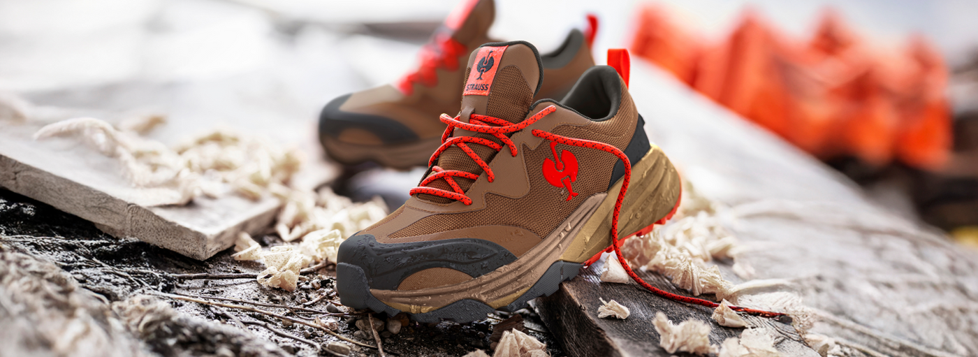 Brown shoes with bright orange laces and logo, resting on a textured surface with wood shavings, against a blurred background.
