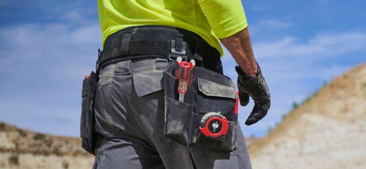 A worker wearing a high-visibility yellow shirt and gray work pants with multiple tool pockets and belt accessories stands outdoors at a worksite.