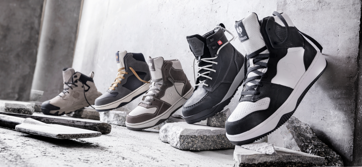 Five work boots and shoes in various styles and colors displayed on concrete blocks against a textured wall.