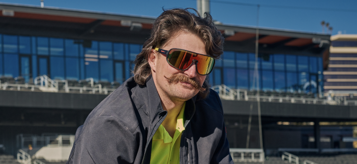 A man wearing reflective safety sunglasses with orange lenses stands outdoors at a construction site on a sunny day.
